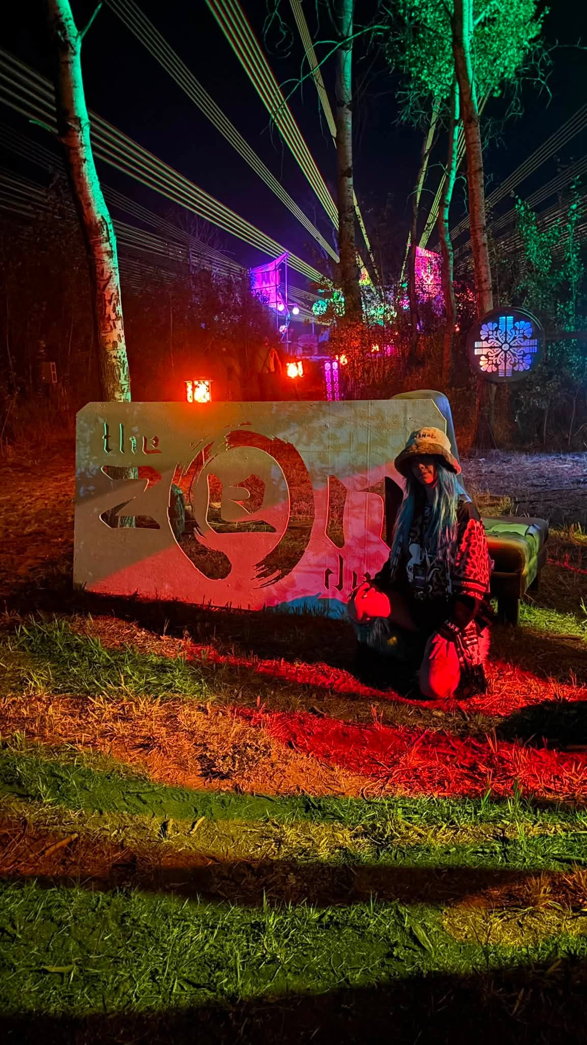 ROZENZA kneeling beside a ZEN sign at a nighttime festival with lasers