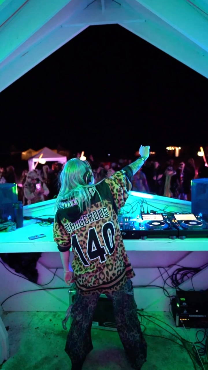 ROZENZA seen from behind on an outdoor festival stage at night, wearing a leopard-print 'DANGEROUS 140' jersey, facing the crowd with the DJ decks in front of her