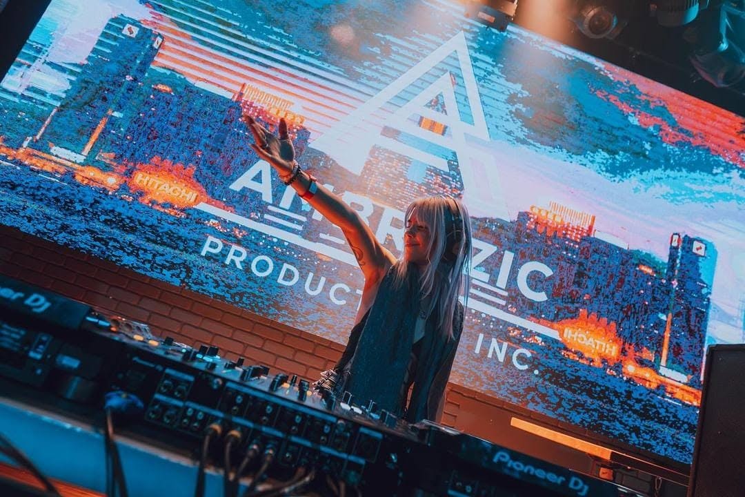 ROZENZA with arm raised at the decks in front of a glowing glitch-art screen